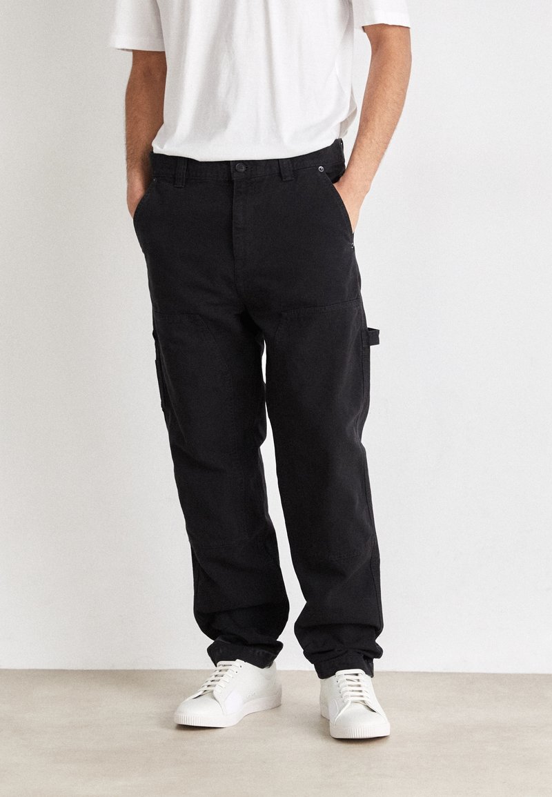 Pantalon cargo noir à coupe décontractée, fermeture à bouton, poches latérales et ourlets retroussés, porté avec des baskets blanches et un t-shirt blanc uni.
