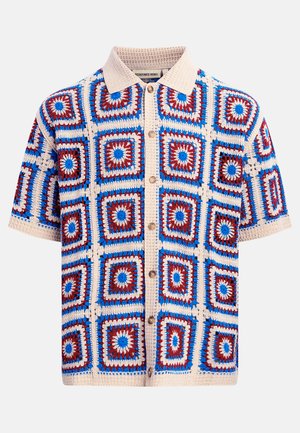 Camisa de manga corta con cuello beige y patrón de crochet con motivos cuadrados en rojo, azul y beige sobre un fondo claro.