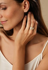 Guldring med texturerade band och en liten ädelsten som accent, bärs på ringfingret. Modellen har medelhudton och långt hår som lätt rör vid hennes ansikte.