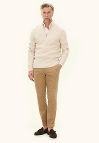 Lichtbeige ritssluitingtrui, witte overhemd eronder, tan corduroybroek en donkerbruine instappers; man staat tegen een lichte achtergrond.