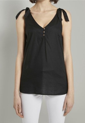 Vrouw draagt een zwarte mouwloze V-hals top met gestrikte schouderbandjes en drie kleine knoopjes, gecombineerd met een witte broek.