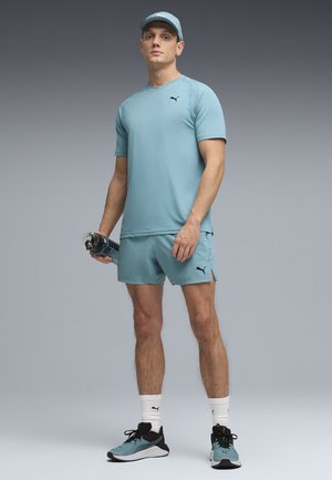 Mann in hellblauem Sportoutfit und Kappe hält eine Wasserflasche und steht vor einem grauen Verlaufshintergrund in Sportkleidung und Turnschuhen.