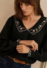 Blusa nera con colletto a scallop e ricami floreali in rosa e blu. Maniche lunghe con dettagli arricciati e una sottile texture.