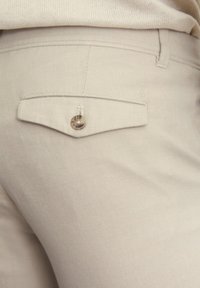 Pantalons beiges en tissu texturé avec une petite poche arrière dotée d'un bouton en métal. Le design comprend un détail de couture.