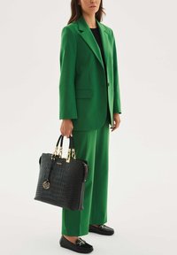 Musta krokotiilinpainokuvioitu käsilaukku, jossa kullanväriset metalliosat, pidetään kirkkaanvihreää pukua ja mustat koristeelliset loafersit pukeutuneen henkilön toimesta.