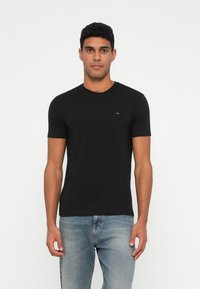 Μαύρη βαμβακερή μπλούζα με κοντά μανίκια και γύρο λαιμό. Διαθέτει ένα μικρό λογότυπο στο στήθος. Συνδυάζεται με ανοιχτό μπλε τζιν παντελόνι.