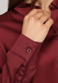 Blouse bordeaux avec une texture lisse, dotée d'un col classique et de poignets à boutons. Les détails remarquables incluent des boutons assortis et des coutures soignées.