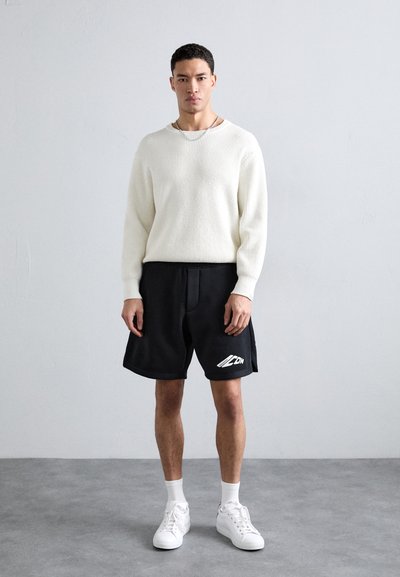 Pull en maille blanche, shorts noirs avec un détail de logo, et baskets blanches. Look athlétique décontracté avec des textures et des couleurs contrastées.