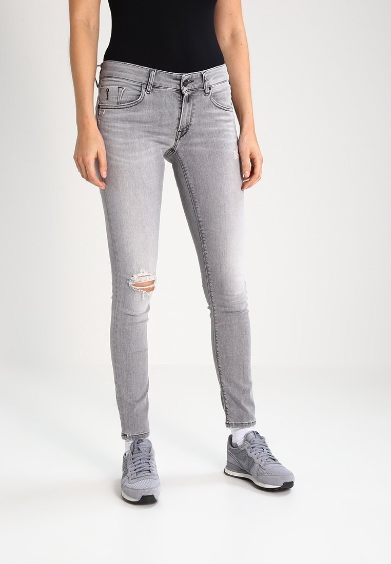 Jeans ajustados en denim gris desgastado con una rodilla desgastada. Combinados con zapatillas grises, mostrando un diseño casual y entallado.