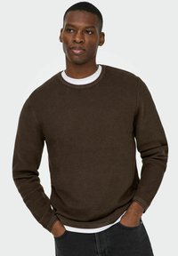 Pull en laine brown texturé avec un col rond, superposé à une chemise blanche. Les manches sont longues avec un détail de poignet côtelé.