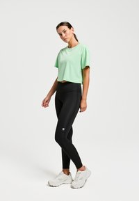 T-shirt de sport à manches courtes, de couleur vert clair et court, associé à un legging noir taille haute avec un logo blanc, et des baskets blanches.
