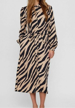 Kobieta ubrana w beżową sukienkę na długi rękaw z czarnymi paskami zebry, zapinaną na guziki, z dopasowanym materiałowym paskiem związanym w talii.