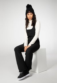 Schwarze Zip-up Latzhose über einem weißen gerippten Rollkragenpullover, kombiniert mit einer gestrickten schwarzen Mütze mit Bommel und schwarzen Sneakers mit weißen Sohlen.