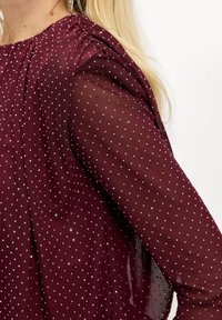 Blouse bordeaux avec manches longues transparentes, ornée de petits pois argentés. Présente des détails froncés aux épaules et est fabriquée en tissu léger.