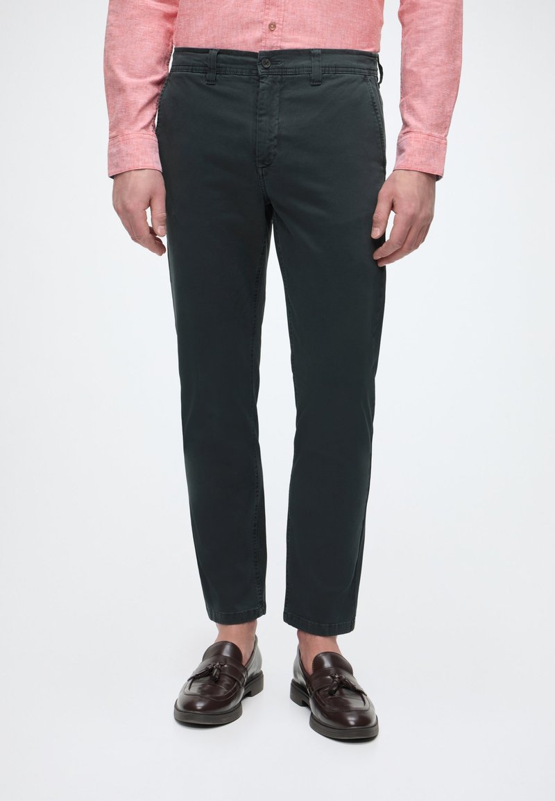 Homme portant un pantalon slim foncé, des mocassins à pampilles marron, et une chemise rose à manches longues avec boutons, debout devant un fond uni.