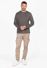 Langärmliges graues Baumwollhemd kombiniert mit beigen Cargo-Hosen. Das Model steht mit entspannten Armen und trägt weiße Sneakers.