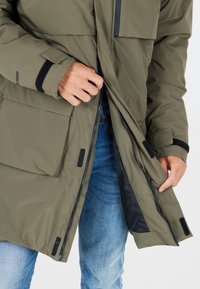 Kaki pufferjacka med dragkedja fram och kardborreaccents. Har en stor ficka på vänster framsidan, med en slät inre foder synlig.