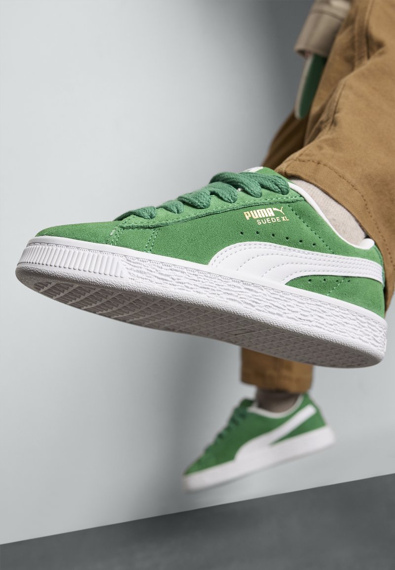 Gröna mockaskor med vita accenter, strukturerad gummisula och snörning. Har Puma-logotyp i guld på sidan.