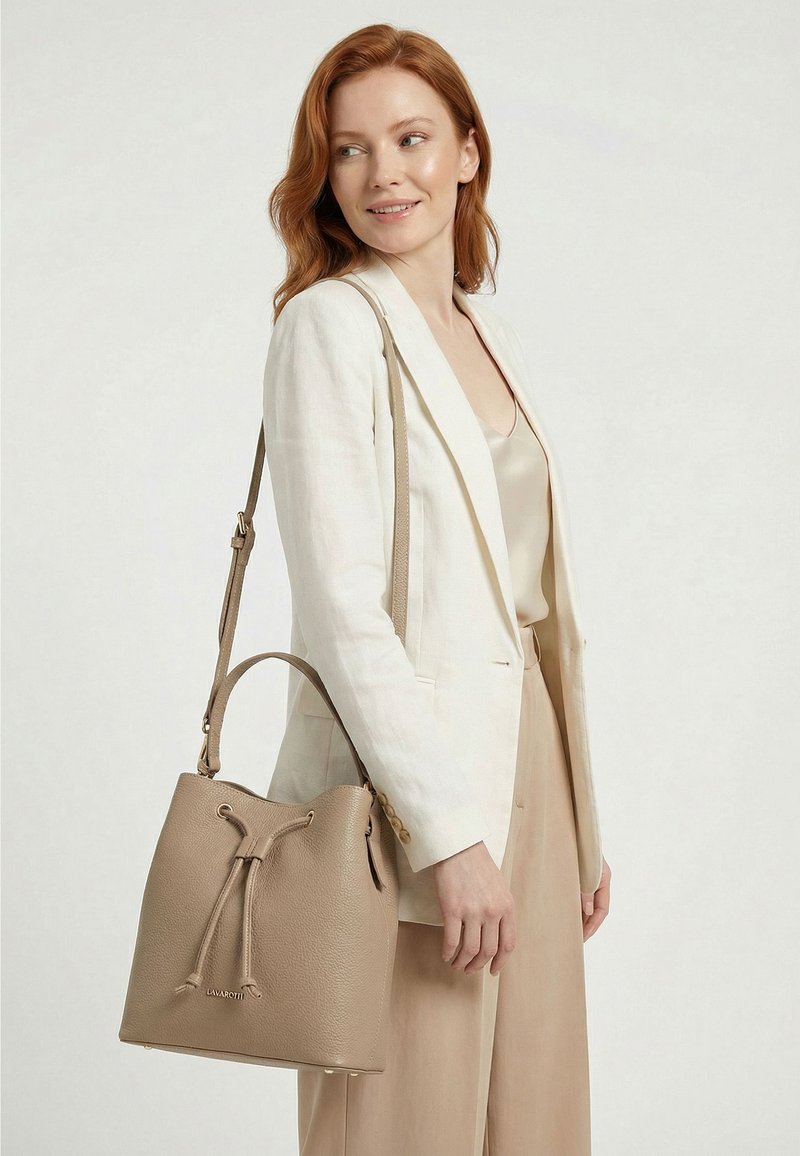Femme rousse portant un blazer blanc et un pantalon beige, portant un sac seau en cuir taupe à cordon sur l'épaule.