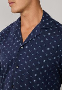 Camisa de manga corta azul marino con un patrón repetido de tijeras en azul claro, con cierre de botones en el frente y cuello con muesca, fabricada en un tejido suave.