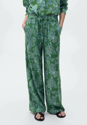 Pantalones verdes de pierna ancha con estampado floral de cachemira azul, cintura elástica, manos en los bolsillos, combinados con sandalias negras de punta abierta sobre fondo claro.