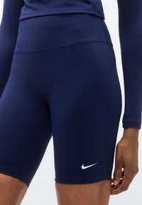 Shorts de sport bleu marine en tissu extensible avec une taille haute, arborant un logo Nike blanc sur la cuisse gauche. Texture lisse.
