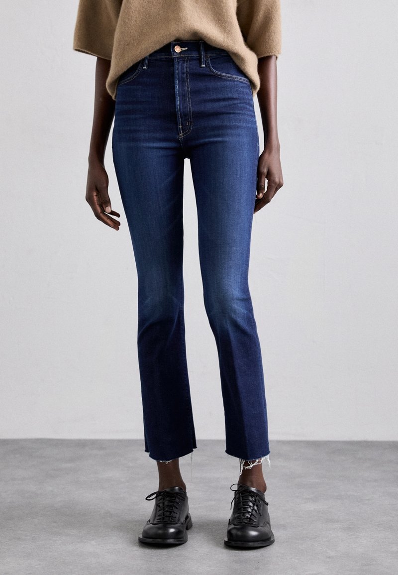 Personne portant un jean court bleu foncé avec des ourlets bruts, un pull beige et des chaussures noires à lacets, debout sur un sol gris.