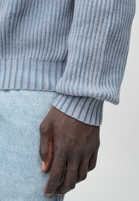 Gaiši zils ribots džemperis ar garām piedurknēm, kas raksturojas ar teksturētu adījumu un kontrastējošām ribotām aprocēm, kombiņots ar gaišiem džinsiem.