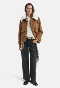 Braune Wolljacke mit einem weißen Schaffellkragen, kombiniert mit einem cremfarbenen Pullover, grauen weiten Jeans und einem karierten Schal. Schwarze Schuhe.
