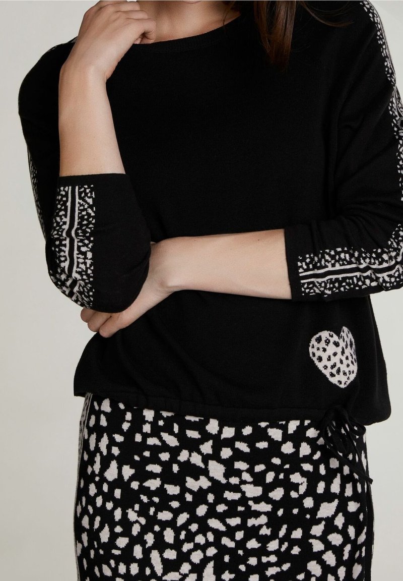 Schwarzer Strickpullover mit gemusterten Ärmeln und einem Herzdetail; kombiniert mit einem schwarz-weiß gesprenkelten Rock. Weiche Textur, lässiges Design.