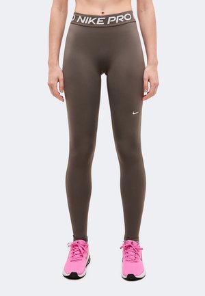 Modèle portant un legging Nike Pro marron foncé avec une taille siglée et des chaussures de running rose vif, sur un fond uni.