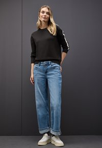 Maglione nero con polsini a costine, jeans in denim azzurro chiaro con vestibilità rilassata e orlo arrotolato, abbinati a sneakers bianche. Design semplice.