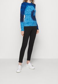 Kvinna i en blå batikmönstrad kabelstickad tröja, svarta leggings och vita sneakers, stående mot en enfärgad vit bakgrund.