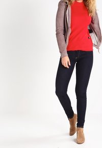 Top de manga larga rojo acanalado, chaqueta de ante gris, jeans oscuros y botas de tobillo beige. La modelo está de pie frente a un fondo claro.