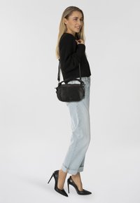 Borsa a tracolla in pelle nera con texture liscia, forma rettangolare, cinturino regolabile e dettaglio della chiusura con zip, indossata da un modello su uno sfondo bianco.