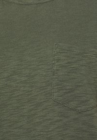 T-shirt vert olive avec une finition texturée, arborant une poche poitrine sur le côté gauche. Col arrondi, manches courtes, design décontracté.