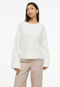 Biały dziany sweter z prążkowanymi mankietami i ściągaczem, o luźnym kroju i okrągłym dekolcie. Noszony na beżowych, teksturowanych spodniach o szerokich nogawkach.