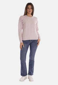 Pullover lavorato a maglia di colore rosa chiaro con scollo a girocollo, indossato con jeans blu e sneakers bianche, con polsini e orlo a costine.