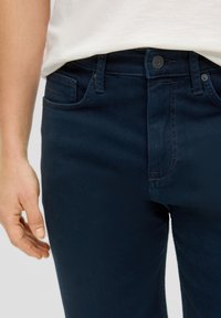Ciemnoniebieskie dżinsy o dopasowanym kroju. Cechy to zapięcie na guzik, pięć kieszeni oraz subtelne detale przeszyć. Gładka faktura.