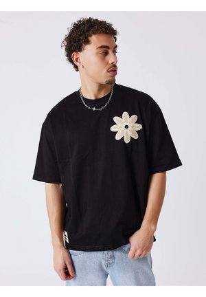 Jonge man met krullend haar die een zwart T-shirt draagt met een grote beige bloemenapplicatie en lichtblauwe spijkerbroek, kijkt zijwaarts.