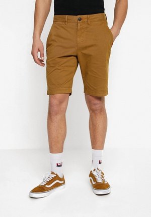 Person trägt senffarbene knielange Shorts, weiße Socken und passende senffarbene Sneakers mit weißen Schnürsenkeln und Sohlen.