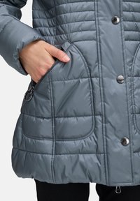 Jasnoszara, pikowana kurtka z ociepleniem, z bocznymi kieszeniami i zatrzaskami. Materiał jest gładki o lekko błyszczącej teksturze.