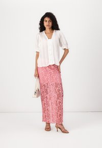 Blouse blanche texturée avec des manches bouffantes et des bords festonnés, associée à une longue jupe en dentelle rose ornée de motifs floraux. Les accessoires incluent un petit sac.