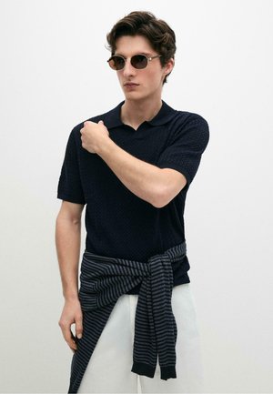 Jonge man met ronde zonnebril, een donker textuur kortärmelig poloshirt en een gestreepte trui die om zijn middel gebonden is, staand tegen een witte achtergrond.