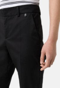 Pantalons noirs ajustés en tissu lisse, avec des poches latérales et un accent en métal argenté arborant un logo "B" sur la taille.