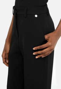 Pantaloni neri con un taglio moderno, texture liscia, tasca laterale e un accento in metallo argentato vicino alla vita.