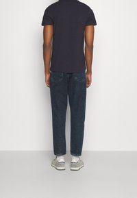 Man står vänd bort ifrån kameran, iförd en svart t-shirt, mörkblå jeans, vita strumpor och grå sneakers med marinblå detaljer.