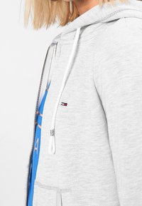 Sudadera gris con capucha y cremallera delantera, cordones blancos y un pequeño parche de logo. Textura de tela suave, bolsillo frontal visible y una camiseta azul debajo.