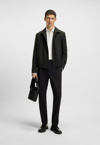 Black zip-up jacket over a white shirt, paired with black pants and black shoes. A small textured black bag is held in the right hand.