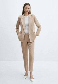 Donna che indossa un completo beige su misura con una maglietta bianca e scarpe a punta bianche, con le mani in tasca contro uno sfondo neutro.